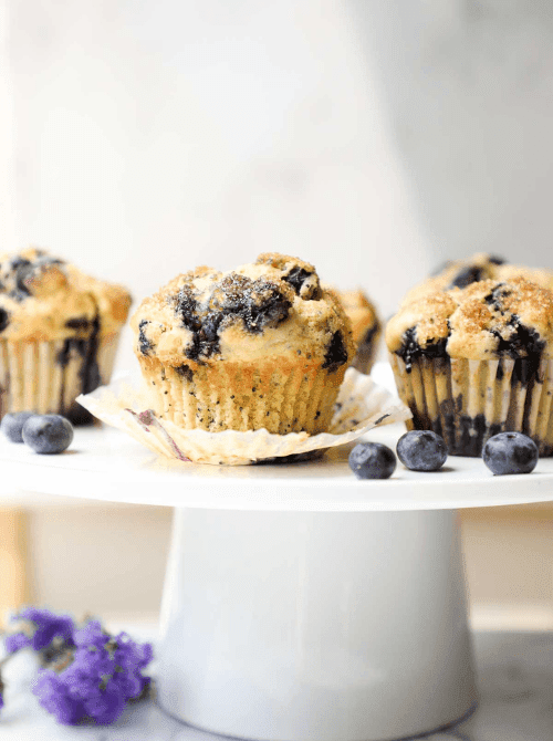 Three tall bakery-style blueberry lemon poppy seed muffins with crackly turbinado sugar tops and unwrapped paper liner displayed on a white cake stand with fresh blueberries and purple flowers at the base — a simple make-ahead Mother's Day brunch idea that bakes in under 25 minutes and looks far more impressive than the effort required.