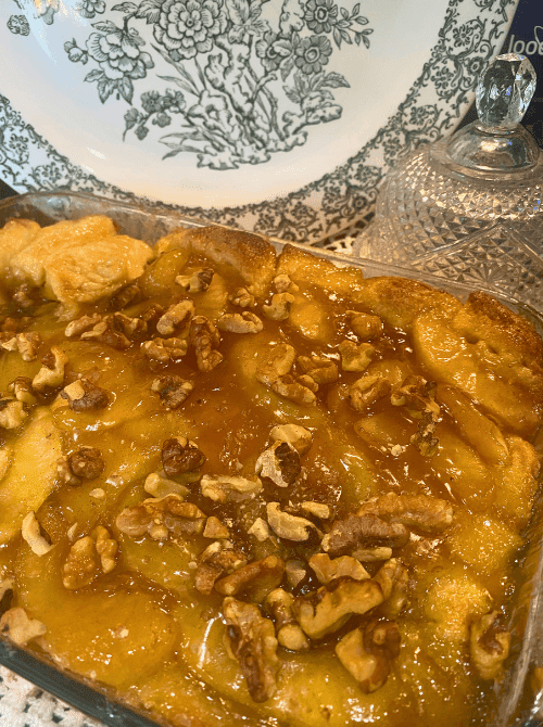 A glass baking dish of caramel apple monkey bread made with canned biscuits, drenched in golden caramel sauce and topped with chopped walnuts, with a vintage green floral plate in the background — an easy make-ahead Mother's Day brunch idea that takes ten minutes to assemble and feeds a crowd for under ten dollars.