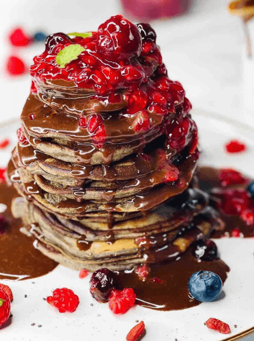 A tall dramatic stack of chocolate berry pancakes drenched in dark chocolate ganache and topped with a glossy mixed berry sauce, surrounded by fresh blueberries, raspberries, and strawberries on a white plate — the showstopping fresh-made addition to a make-ahead Mother's Day brunch ideas spread that fills the kitchen with the smell that tells everyone the celebration has started.