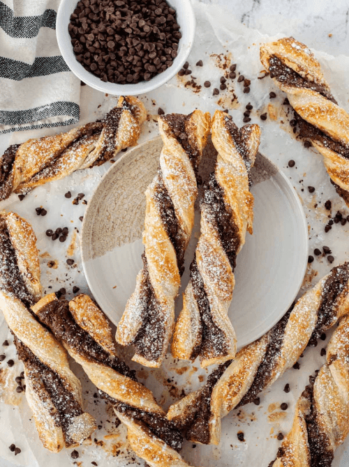 Flaky puff pastry chocolate twist pastries filled with Nutella and mini chocolate chips, dusted with powdered sugar and arranged around a small plate on parchment paper with a bowl of chocolate chips — a bakery-style make-ahead Mother's Day brunch idea that comes together in under 25 minutes with store-bought puff pastry.