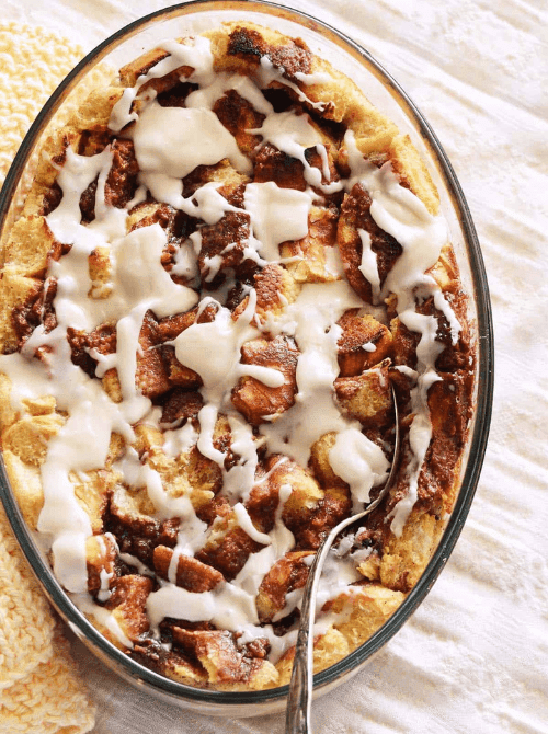 An oval glass baking dish of golden brioche French toast casserole drizzled with cream cheese glaze and a serving spoon resting inside, on a white linen surface — a rich and custardy make-ahead Mother's Day brunch idea that soaks overnight and feeds ten to twelve people with almost no morning effort.