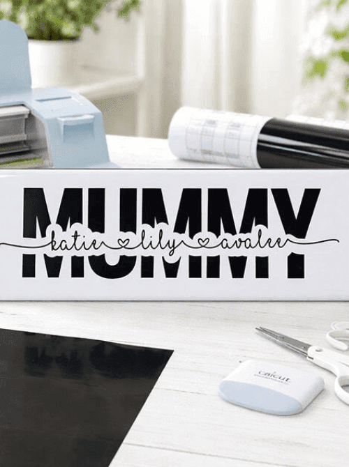 Personalized Cricut knockout tile with bold MUMMY text and children's names in script vinyl on a white ceramic tile, with a Cricut machine and vinyl roll in the background — a custom handmade keepsake for DIY Mother's Day gift baskets.