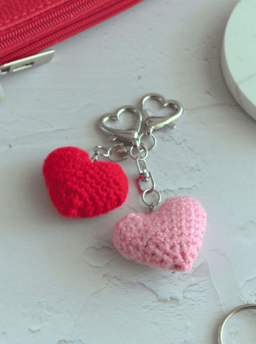 Two small crocheted heart keychains in red and pink attached to a silver heart-shaped clasp — a quick handmade accessory gift idea for DIY Mother's Day gift baskets.