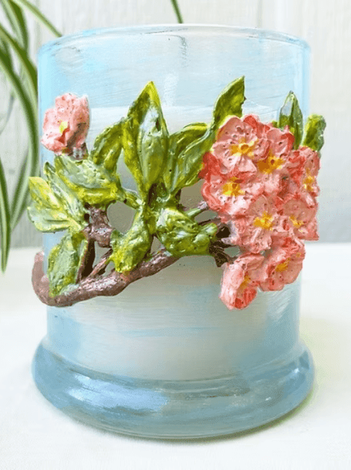 A DIY glass candle holder painted in soft blue with dimensional pink floral and green leaf clay embellishments — a handmade home décor gift idea for DIY Mother's Day gift baskets.