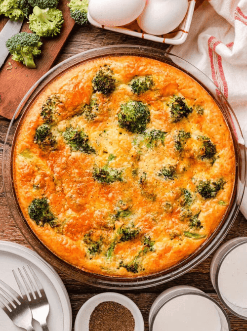 Golden crustless ham and broccoli quiche baked in a glass pie dish with visible broccoli florets throughout, surrounded by fresh eggs, milk, and plates on a wooden surface — a simple make-ahead Mother's Day brunch idea that comes together in five minutes and bakes while you sleep.