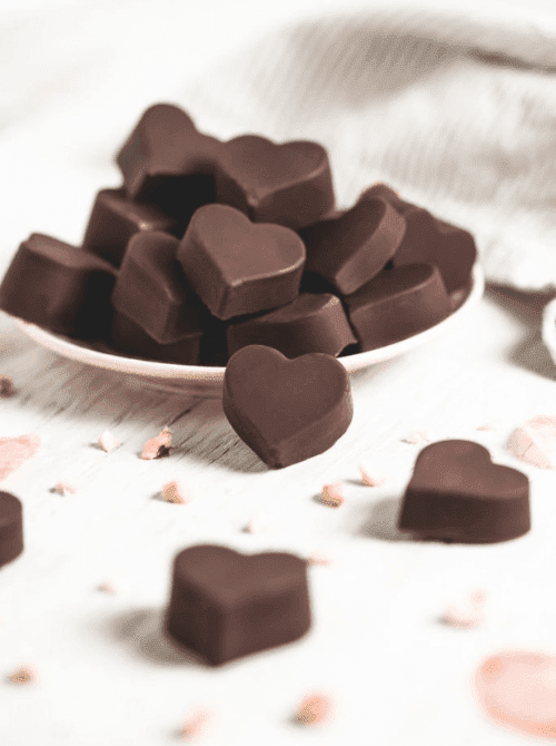 Heart-shaped healthy hazelnut chocolate truffles piled on a white plate with scattered hazelnuts — a refined-sugar-free homemade chocolate gift idea for DIY Mother's Day gift baskets.