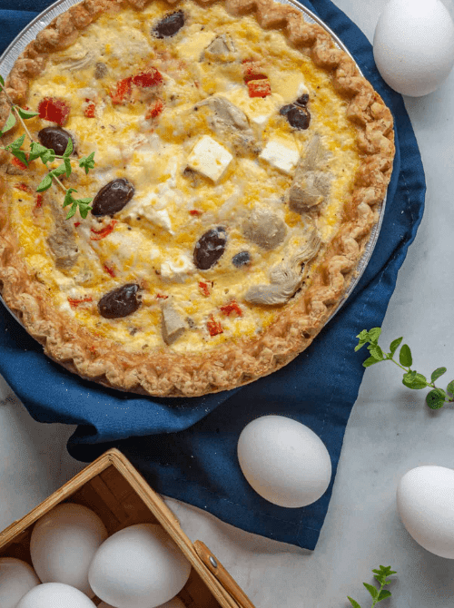 Golden flaky-crusted quiche filled with artichokes, kalamata olives, roasted red peppers, and cubed feta cheese on a blue linen with fresh eggs on a marble surface — a Mediterranean-inspired make-ahead Mother's Day brunch idea that tastes even better served at room temperature.