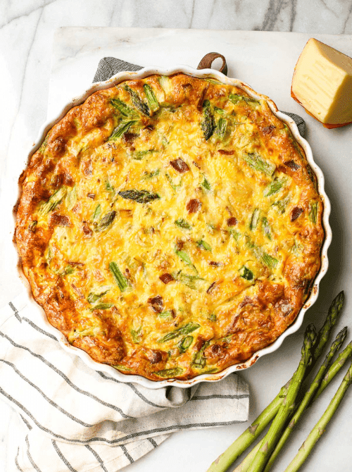Golden baked leek and bacon frittata with asparagus and melted fontina cheese in a white ceramic pie dish, served on a marble surface — a make-ahead Mother's Day brunch idea that goes from fridge to oven with no morning stress.