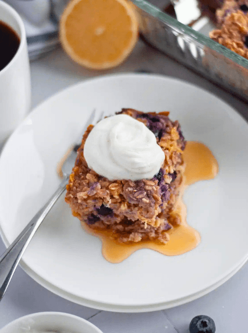 A square of lemon blueberry baked oatmeal topped with a dollop of whipped cream and drizzled with maple syrup on a white plate, with a halved lemon, coffee, and baking dish in the background — a light and wholesome make-ahead Mother's Day brunch idea that is naturally gluten-free and ready to reheat in minutes.