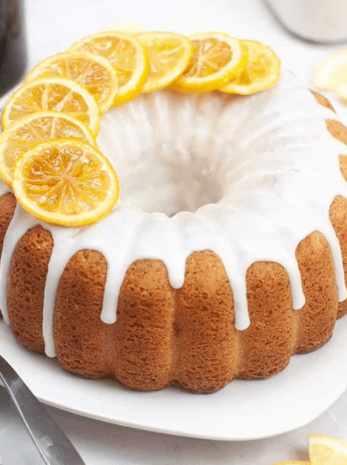 A golden lemon Nothing Bundt cake copycat drizzled with white lemon glaze and topped with candied lemon slices arranged in a ring on a white cake stand — a stunning make-ahead Mother's Day brunch idea that bakes the day before, frosts the morning of, and serves ten to twelve guests straight from the stand.