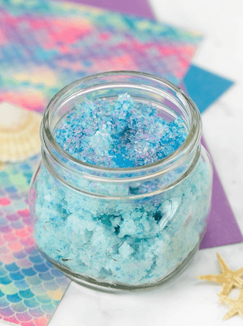 Blue and purple swirled mermaid sugar scrub in a glass jar with a starfish and mermaid-print paper in the background — a fun and colorful homemade bath gift for DIY Mother's Day gift baskets.