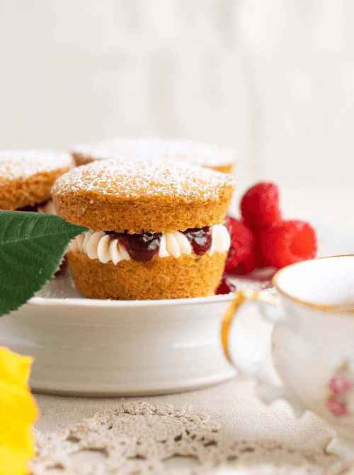 A mini Victoria sponge cake dusted with powdered sugar and filled with vanilla buttercream and raspberry jam displayed on a small white cake stand, with fresh raspberries, a gold-rimmed teacup, and more mini cakes softly blurred in the background — an elegant make-ahead Mother's Day brunch idea baked in a muffin tin that looks like it came from a British tea room.