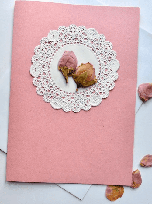 6:40 PMA handmade pressed flower card with dried pink rosebuds on a white paper doily centered on a pink cardstock card with a white envelope — a beautiful homemade keepsake for DIY Mother's Day gift baskets.