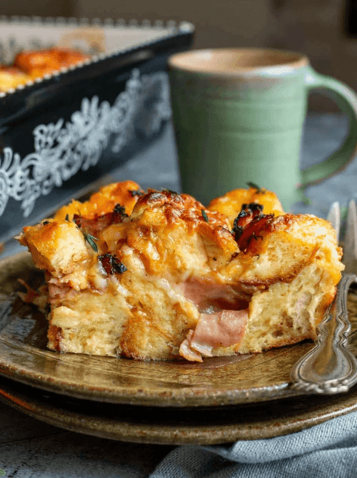 A golden slice of rich bread and butter pudding with layers of ham and melted cheese served on a rustic green plate with a coffee mug and baking dish in the background — a hearty make-ahead Mother's Day brunch idea that assembles the night before and feeds a crowd with almost no morning effort.