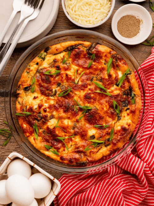 Golden baked spinach mushroom feta quiche topped with sliced green onions in a glass pie dish, surrounded by eggs and shredded cheese on a wooden surface — an easy make-ahead Mother's Day brunch idea that can be assembled the night before and baked fresh in the morning.