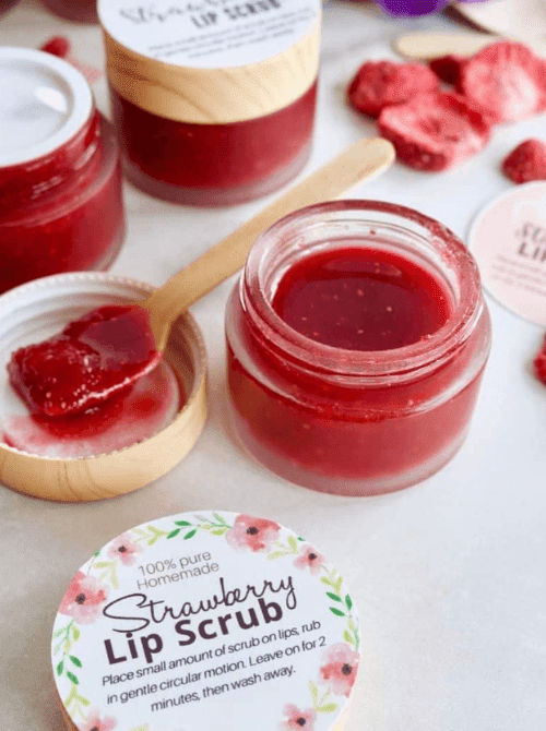 Small glass jars of homemade strawberry lip scrub with wooden spoons and a floral printable label, styled with freeze-dried strawberries — a handmade beauty gift idea for DIY Mother's Day gift baskets.