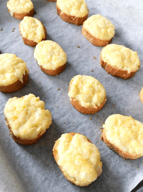 Rows of warm egg and cheese crostini topped with a golden grated egg and cheese mixture on toasted baguette slices arranged on parchment paper — a bite-sized make-ahead Mother's Day brunch idea that preps in minutes and bakes fresh in the morning.