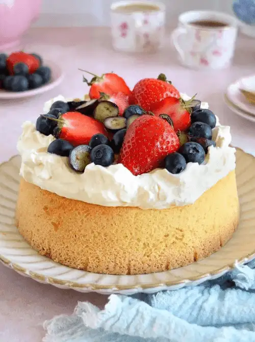 Air fryer butter-free genoise sponge cake topped with whipped cream and fresh strawberries and blueberries on a vintage cake plate — a light homemade strawberry cake recipe made entirely in an air fryer
