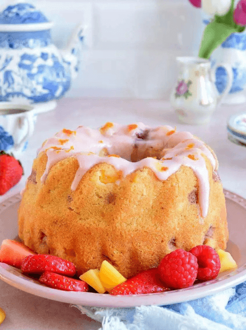 Air fryer mango strawberry bundt cake with pink strawberry glaze dripping down the ridged sides on a pink plate, surrounded by fresh strawberries and mango chunks — a visually stunning homemade strawberry cake recipe made in an air fryer