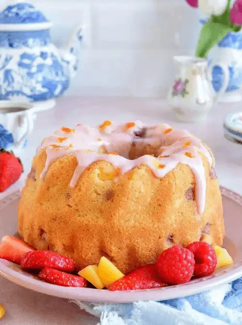 Air fryer mango strawberry bundt cake with pink strawberry glaze dripping down the ridged sides on a pink plate, surrounded by fresh strawberries and mango chunks — a visually stunning homemade strawberry cake recipe made in an air fryer