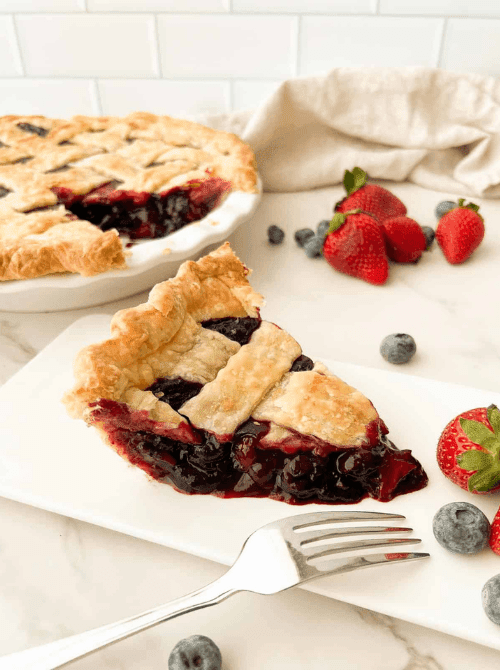 A slice of blueberry strawberry pie with a flaky golden lattice crust and deep purple jammy berry filling on a white plate, with the full pie and fresh berries visible in the background — a beautiful summer strawberry dessert recipe that doubles as a red white and blue crowd-pleaser.