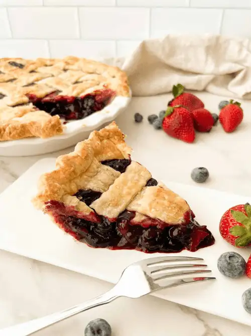 A slice of blueberry strawberry pie with a flaky golden lattice crust and deep purple jammy berry filling on a white plate, with the full pie and fresh berries visible in the background — a beautiful summer strawberry dessert recipe that doubles as a red white and blue crowd-pleaser.