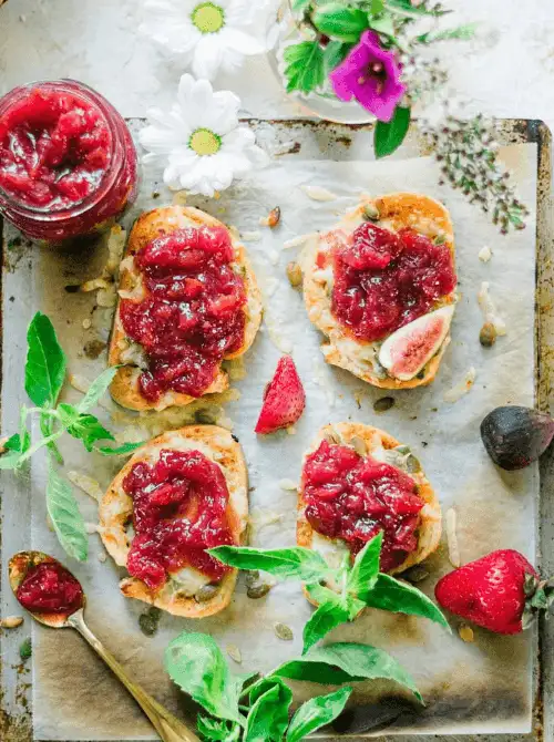 Overhead view of toasted crostini topped with melted cheese and strawberry fig preserves on a rustic baking sheet with fresh herbs and flowers — a savory-sweet fresh strawberry brunch recipe ready in 15 minutes.
