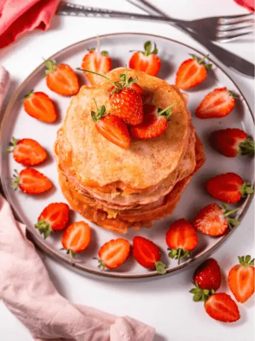 Stack of fluffy strawberry pancakes on a gray plate surrounded by fresh halved strawberries — an easy fresh strawberry brunch recipe for a crowd.