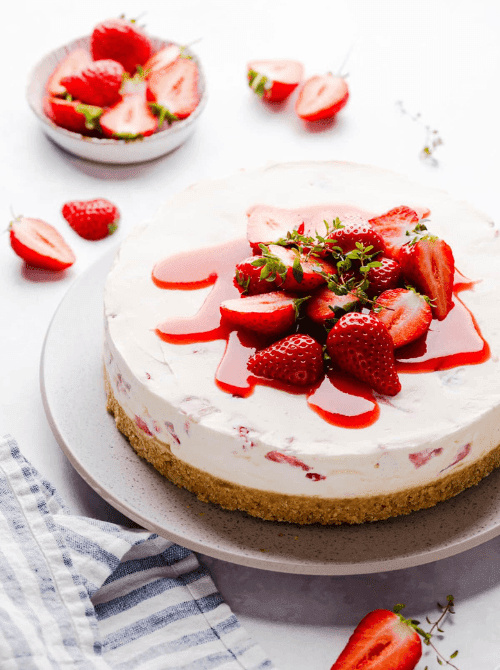 A whole no-bake strawberry white chocolate cheesecake on a grey serving plate topped with fresh sliced strawberries and a glossy strawberry sauce, with scatter…A whole no-bake strawberry white chocolate cheesecake on a grey serving plate topped with fresh sliced strawberries and a glossy strawberry sauce, with scattered berries and a striped linen nearby — an elegant summer strawberry dessert that requires zero oven time.