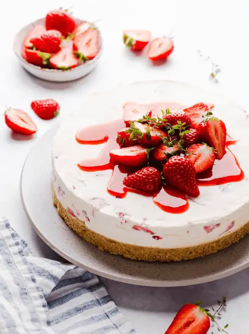 A whole no-bake strawberry white chocolate cheesecake on a grey serving plate topped with fresh sliced strawberries and a glossy strawberry sauce, with scatter…A whole no-bake strawberry white chocolate cheesecake on a grey serving plate topped with fresh sliced strawberries and a glossy strawberry sauce, with scattered berries and a striped linen nearby — an elegant summer strawberry dessert that requires zero oven time.