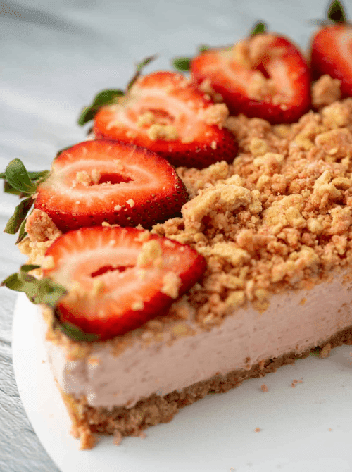 A close-up of a no-bake strawberry cheesecake with a golden graham cracker crust, creamy pink filling, crumble topping, and halved fresh strawberries fanned across the top — one of the most crowd-pleasing things to make with strawberries all summer.