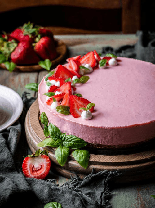 Pink frosted cake on a wooden stand, topped with sliced strawberries and small white decorations, with fresh basil leaves nearby.