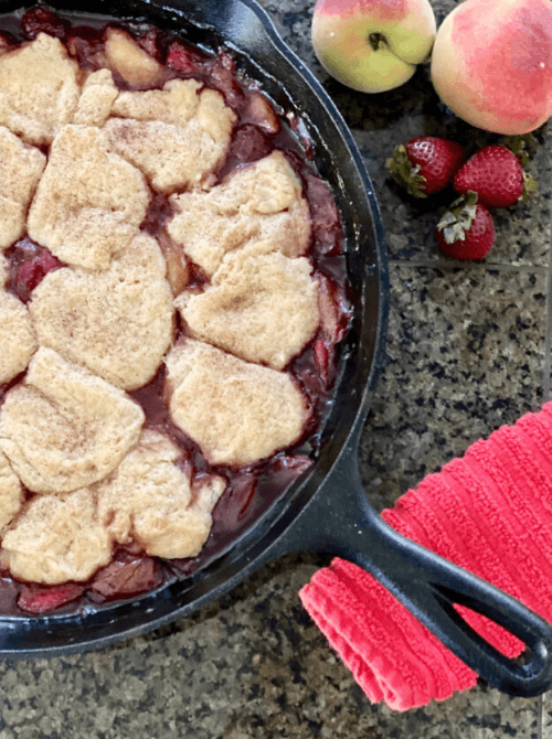 A cast iron skillet filled with old fashioned peach strawberry cobbler topped with golden biscuit rounds, with fresh peaches and strawberries on the side — a hearty summer strawberry dessert recipe that feeds a crowd.