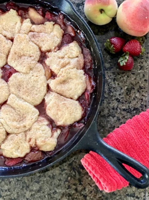 A cast iron skillet filled with old fashioned peach strawberry cobbler topped with golden biscuit rounds, with fresh peaches and strawberries on the side — a hearty summer strawberry dessert recipe that feeds a crowd.
