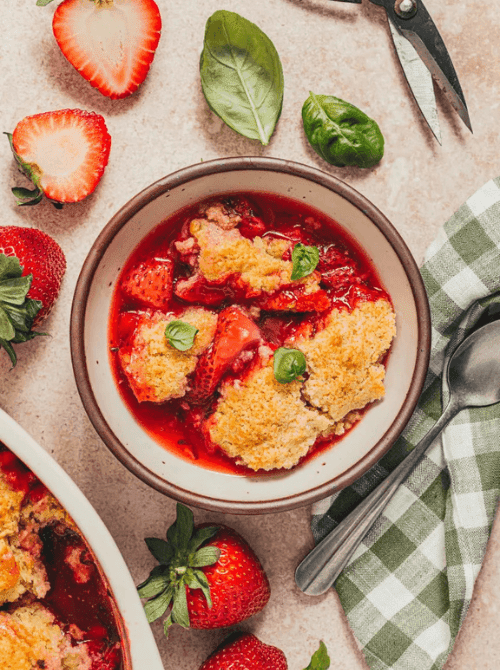 A bowl of gluten-free strawberry basil cobbler with golden biscuit topping and fresh basil leaves, surrounded by halved strawberries and a green checked napkin — a unique twist on summer strawberry desserts.