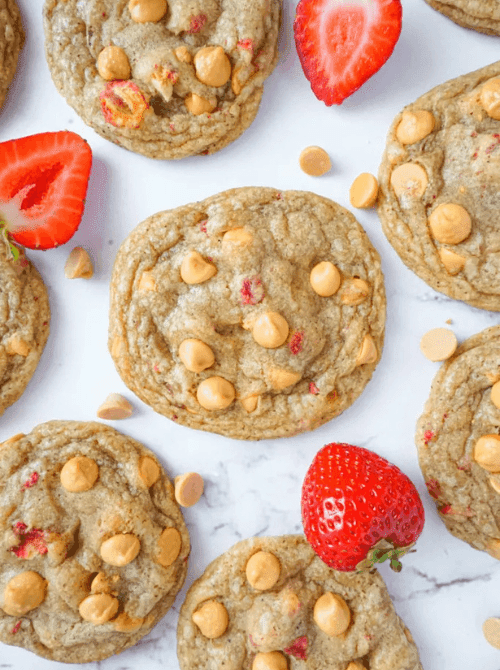 Overhead view of strawberry butterscotch cookies loaded with butterscotch chips and freeze-dried strawberry pieces, scattered on a marble surface with fresh strawberries — a unexpected and delicious thing to make with strawberries.