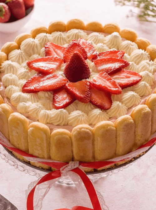 Strawberry Charlotte cake on a glass cake stand with ladyfinger border and fresh sliced strawberries on top — one of the most beautiful no-bake strawberry cake recipes you can make at home.