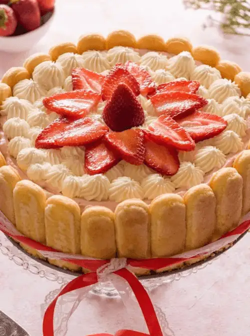 Strawberry Charlotte cake on a glass cake stand with ladyfinger border and fresh sliced strawberries on top — one of the most beautiful no-bake strawberry cake recipes you can make at home.
