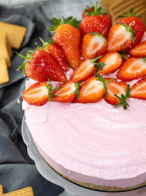 A whole no-bake strawberry cheesecake on a grey cake stand with a fluffy pink cream cheese filling and a mound of fresh whole and halved strawberries piled on top, with graham crackers visible in the background — one of the most impressive things to make with strawberries when you need a dessert that feeds a crowd.