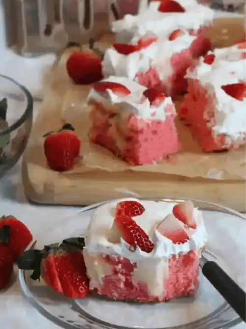 Slice of strawberry cheesecake poke cake on a glass plate showing pink cheesecake pudding soaked through the cake with whipped cream and fresh strawberries on top — an easy homemade strawberry cake recipe that feeds a crowd