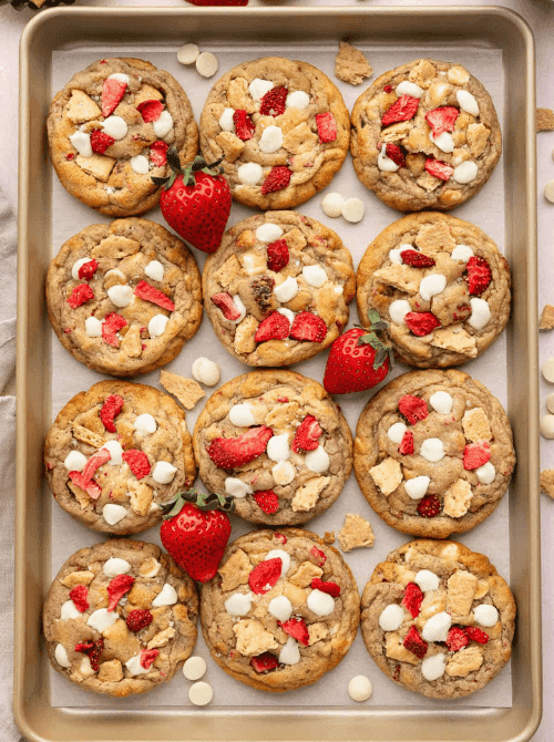 A gold baking sheet lined with strawberry cheesecake cookies studded with white chocolate chips, graham cracker pieces, and freeze-dried strawberries — a fun and creative strawberry dessert recipe that tastes like cheesecake in cookie form.