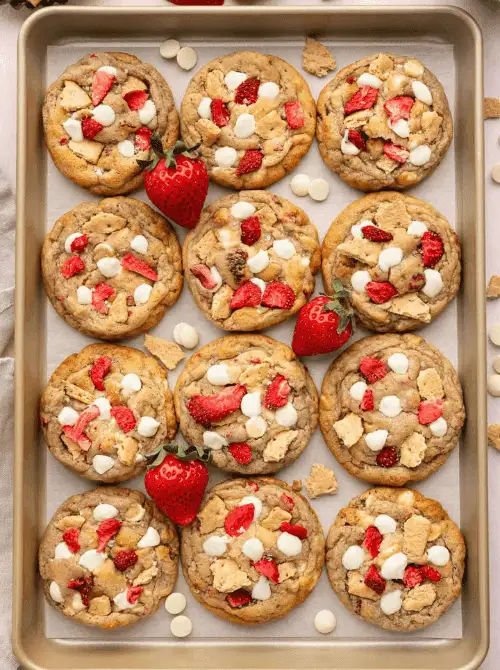 A gold baking sheet lined with strawberry cheesecake cookies studded with white chocolate chips, graham cracker pieces, and freeze-dried strawberries — a fun and creative strawberry dessert recipe that tastes like cheesecake in cookie form.