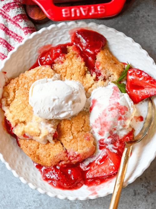 A serving of strawberry cobbler in a white scalloped bowl topped with two scoops of vanilla ice cream, with a gold spoon and a red Staub skillet in the background — one of the easiest summer strawberry desserts you can make.