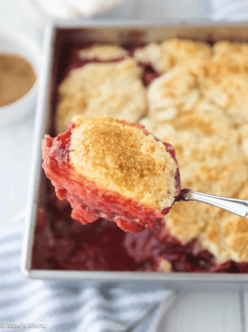 A spoonful of strawberry cobbler held up close showing the golden biscuit topping and jammy red berry filling, with the full baking pan visible in the background — a classic strawberry dessert recipe perfect for summer.