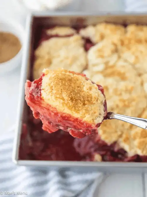 A spoonful of strawberry cobbler held up close showing the golden biscuit topping and jammy red berry filling, with the full baking pan visible in the background — a classic strawberry dessert recipe perfect for summer.