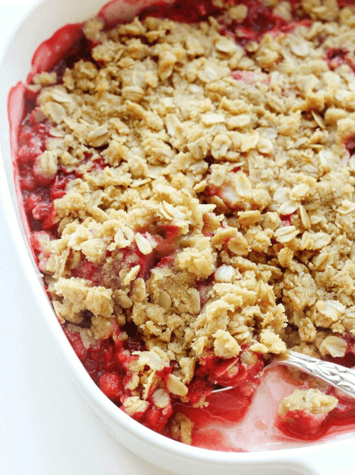A white oval baking dish of strawberry rhubarb crisp with a thick golden oat topping and bubbling red berry filling, with a spoon resting in the corner — a simple strawberry dessert recipe that lets the fruit do the work.