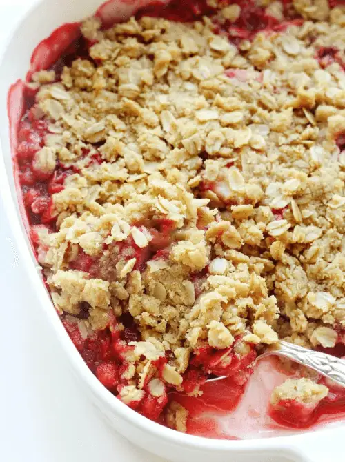 A white oval baking dish of strawberry rhubarb crisp with a thick golden oat topping and bubbling red berry filling, with a spoon resting in the corner — a simple strawberry dessert recipe that lets the fruit do the work.