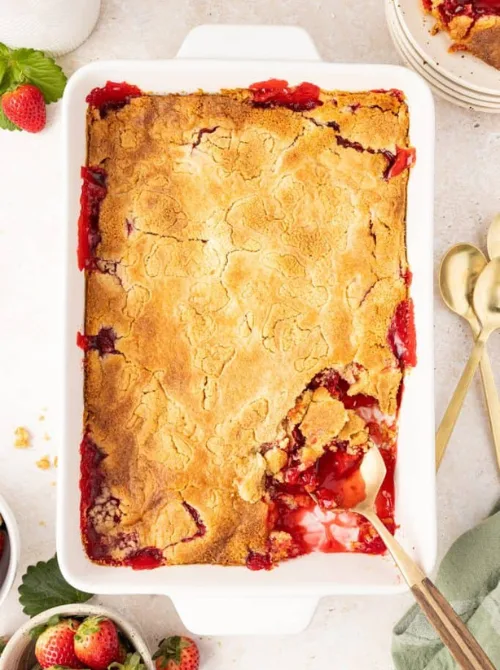 Overhead view of strawberry dump cake in a white baking dish with a golden crust and bubbling strawberry filling, spoon scooping out the first serving — the easiest three-ingredient strawberry cake recipe for a crowd