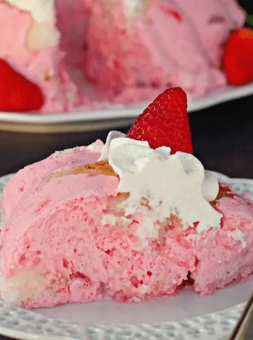 Close-up of a slice of strawberry Jello angel food cake with pink strawberry cream filling soaked through the torn cake, topped with whipped cream and a fresh strawberry — a retro no-bake strawberry cake recipe that tastes better than it looks