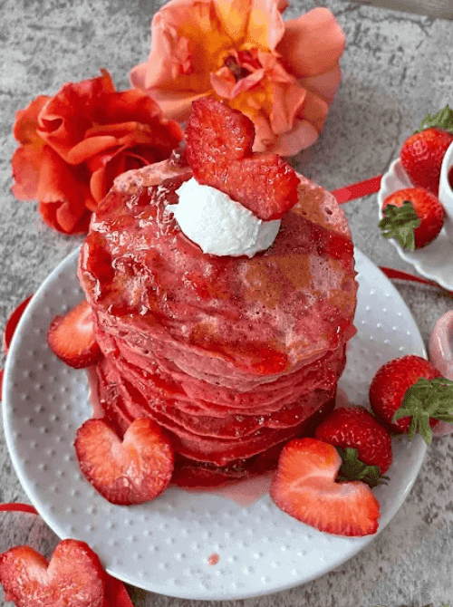 Tall stack of strawberry ombre pancakes in graduating shades of pink, topped with whipped cream and fresh sliced strawberries — a stunning fresh strawberry brunch recipe.